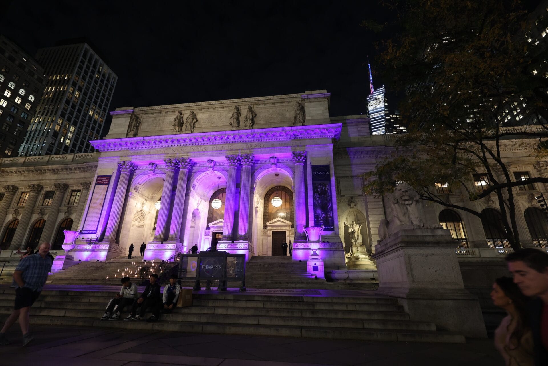 New York Public Library facade at night lit in purple, with grand steps, columns, and statues; people seated on the steps.