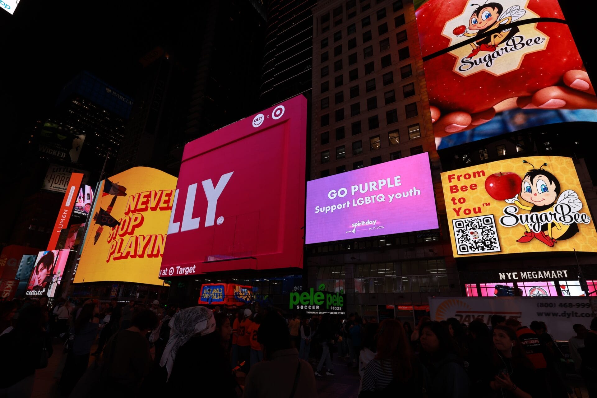 Nighttime Times Square scene with large colorful LED billboards, including pink, purple, and SugarBee ads, and a crowd of pedestrians below.