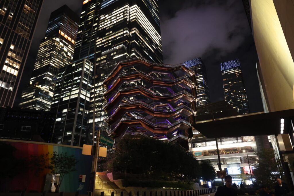 Night view of tall glass skyscrapers at Hudson Yards with The Vessel spiraling copper stairs centerpiece in foreground.